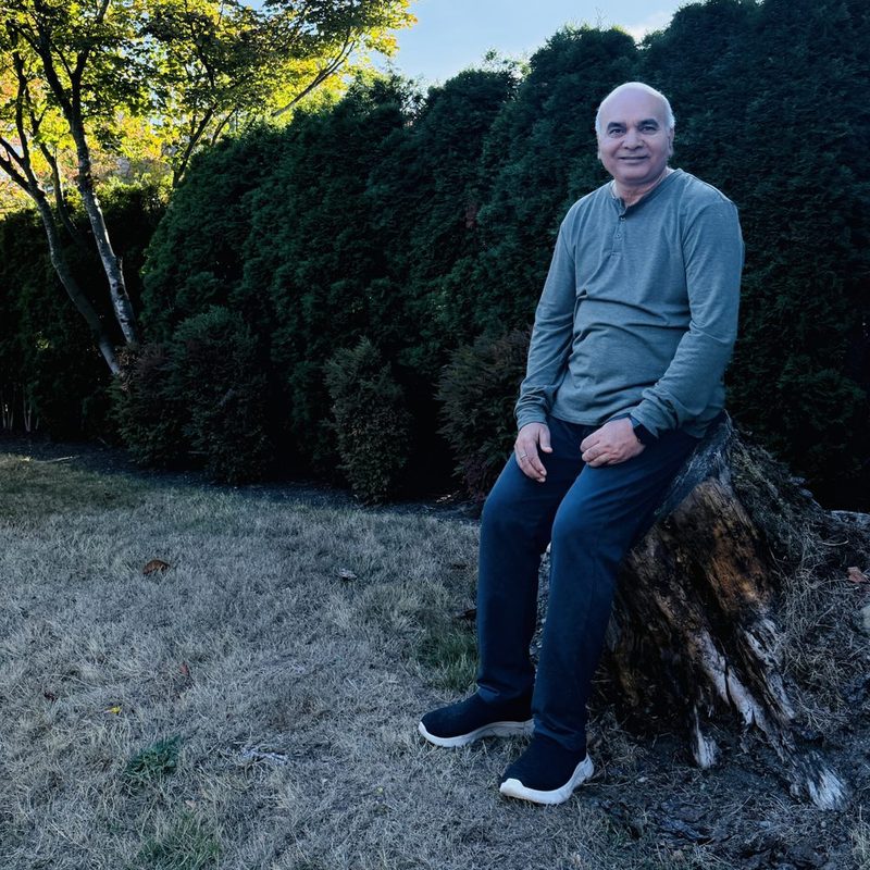 Dr. Dwivedi seated on a tree stump in a garden, with a tall hedge behind him